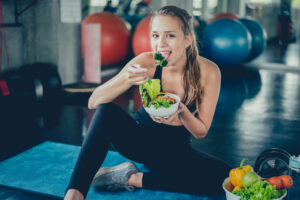 Young woman in sportswear sitting on a gym mat, eating a fresh salad, with colorful vegetables and fitness equipment in the background.